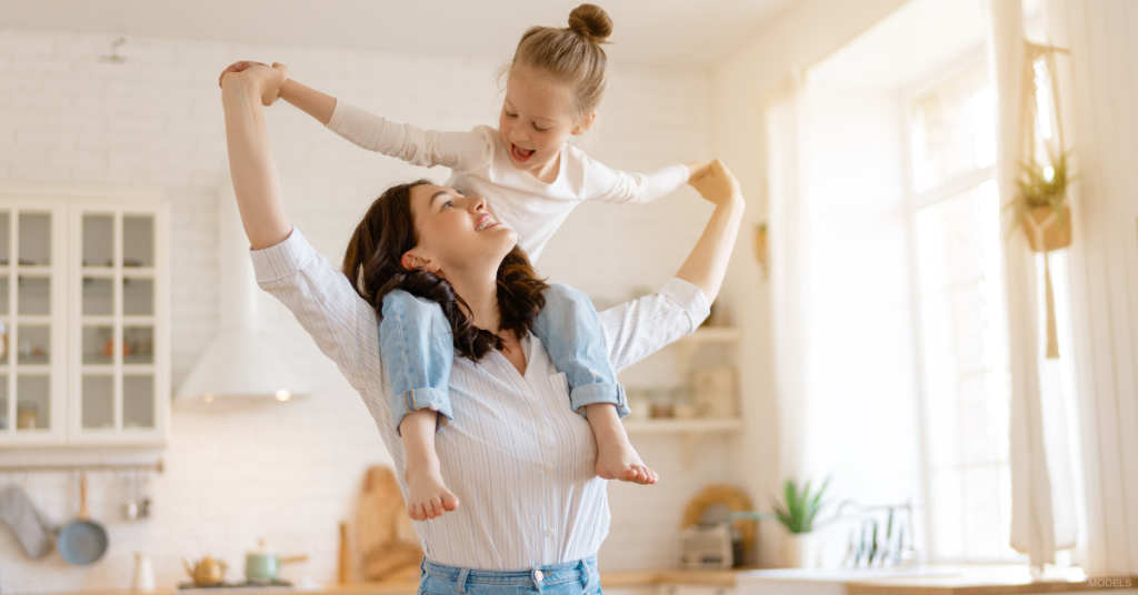 A mother with her daughter on her shoulders in the kitchen (models)