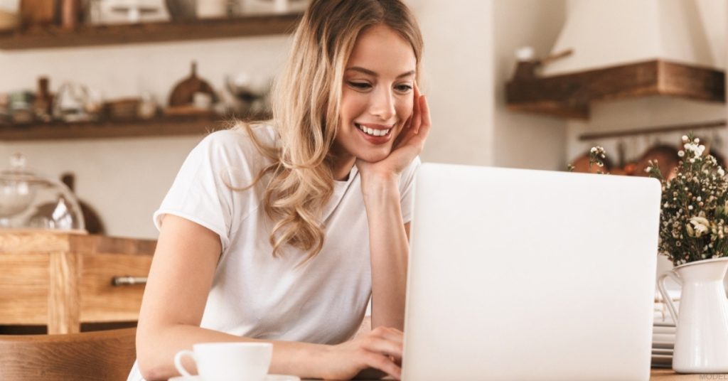 Woman smiles while sitting at a table with a laptop (MODEL)