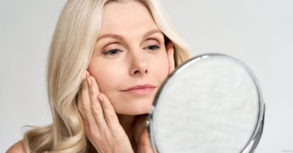 A mature woman gently touches her face while looking in the mirror (MODEL)